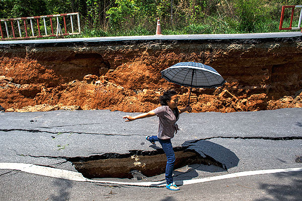 Женщина с зонтиком прыгает через яму в разрушенной землетрясением дороге.