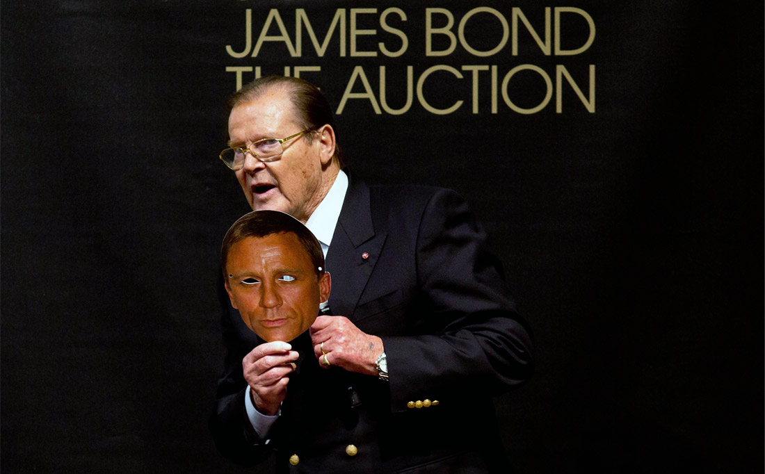 Актер Роджер Мур держит в руках маску с изображением Дэниэла Крэйга на аукционе, посвященном 50-летнему юбилею Бондианы