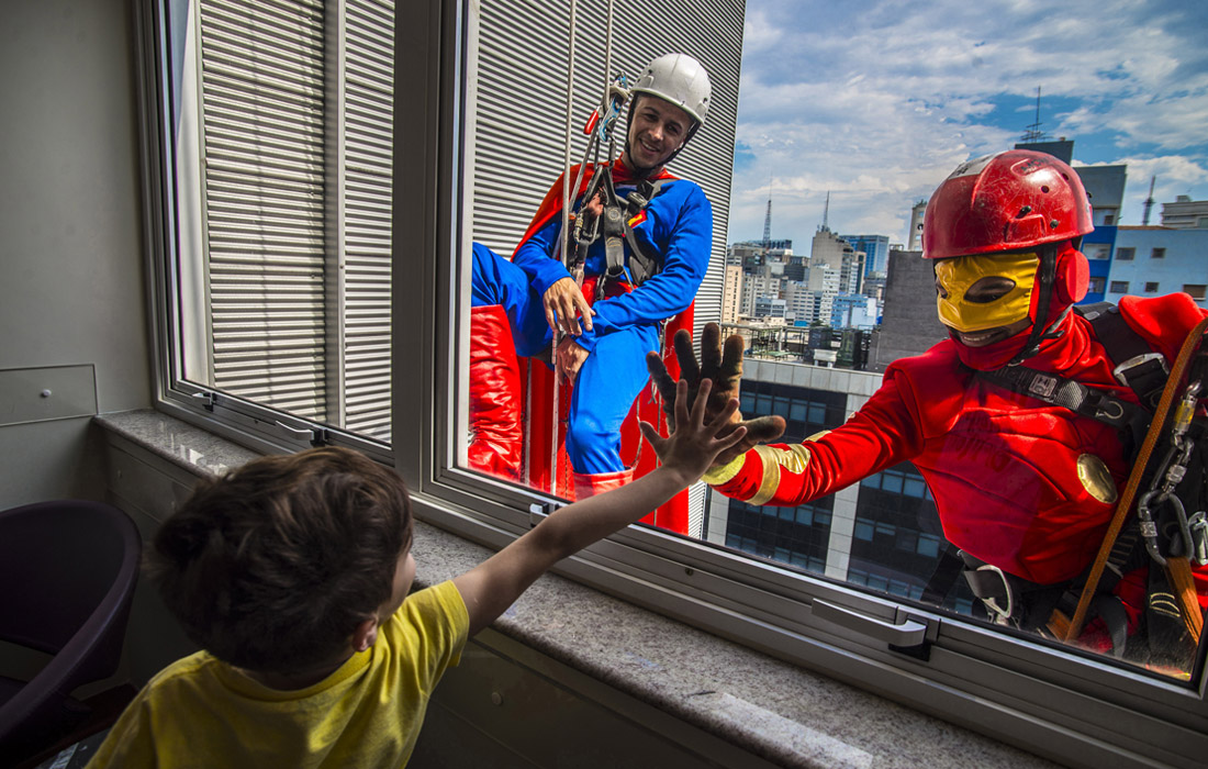 Юный пациент приветствует переодетых в супергероев мойщиков окон в больнице Сан-Паулу. Бразилия.