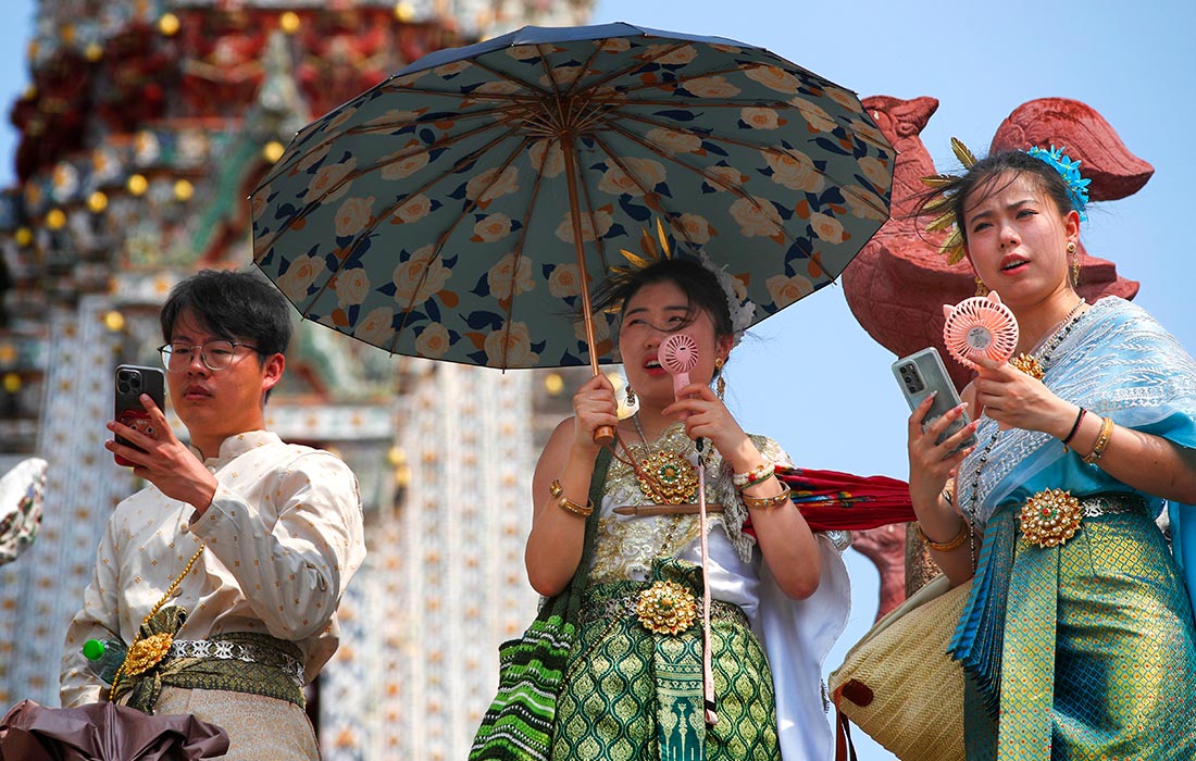 Китайские туристы, одетые в традиционные тайские костюмы, охлаждаются с помощью ручных вентиляторов у Храма Зари в Бангкоке